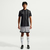 Ein Mann in einem schwarzen Streifen-Trikot und grauen Shorts steht vor weißem Hintergrund.
