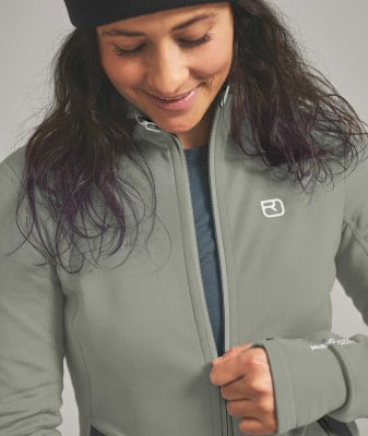 Eine Frau schließt eine graue Jacke mit weißem Logo und lila Haarsträhnen. Eine Frau schließt eine graue Jacke mit weißem Logo und lila Haarsträhnen.