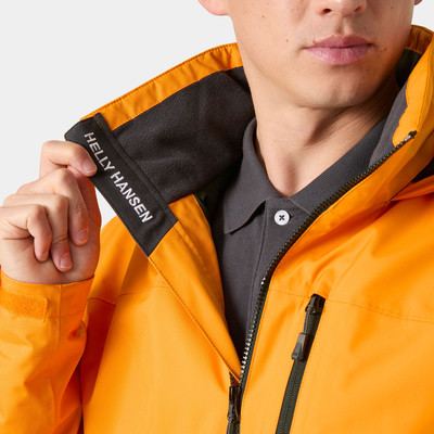 Egy férfi Helly Hansen feliratú narancssárga dzseki gallérját igazgatja. Egy férfi Helly Hansen feliratú narancssárga dzseki gallérját igazgatja.