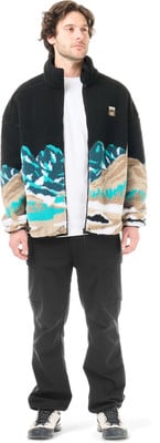 Ein Mann in schwarzer Hose und einer Fleecejacke mit Bergmotiv. Ein Mann in schwarzer Hose und einer Fleecejacke mit Bergmotiv.