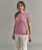 Eine Frau in einem rosa T-Shirt und hellen Hosen posiert vor einem grauen Hintergrund.