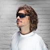 Mann mit lockigem Haar und Sonnenbrille vor weißer Ziegelwand.