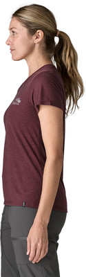 Eine Frau in einem T-Shirt und einer Hose posiert im Profil vor weißem Hintergrund.
