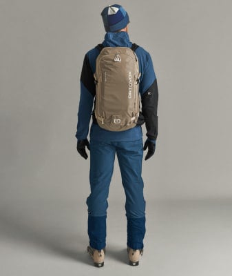 Ein Mann in Skibekleidung mit Rucksack auf dem Rücken steht vor grauem Hintergrund. Ein Mann in Skibekleidung mit Rucksack auf dem Rücken steht vor grauem Hintergrund.