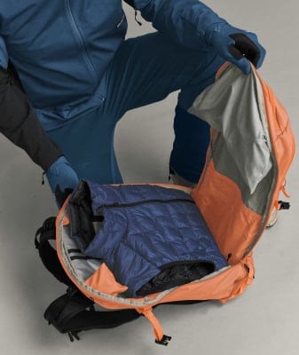 Eine Person in blauer Kleidung packt eine Daunenjacke in einen orangefarbenen Rucksack. Eine Person in blauer Kleidung packt eine Daunenjacke in einen orangefarbenen Rucksack.