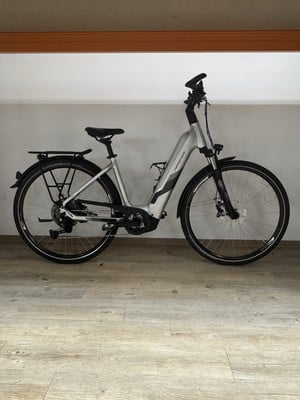 Ein silbernes E-Bike steht auf einem Holzboden in einem Raum. Ein silbernes E-Bike steht auf einem Holzboden in einem Raum.