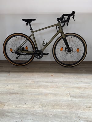 Ein grünes Gravelbike mit schwarzen Felgen und braunen Reifen steht auf einem Holzboden. Ein grünes Gravelbike mit schwarzen Felgen und braunen Reifen steht auf einem Holzboden.