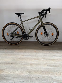 Ein grünes Gravelbike mit schwarzen Felgen und braunen Reifen steht auf einem Holzboden.