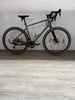 Ein grünes Gravelbike mit schwarzen Felgen und braunen Reifen steht auf einem Holzboden.