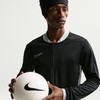 Ein Mann hält einen Fußball und trägt eine schwarze Nike-Jacke.