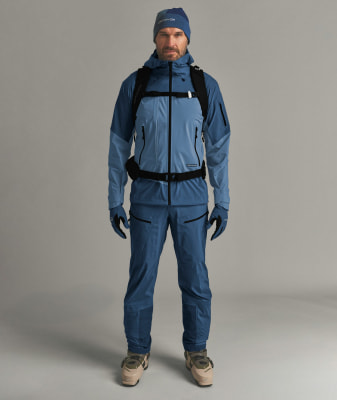Ein Mann in blauer Skibekleidung und Skischuhen steht vor einem grauen Hintergrund.