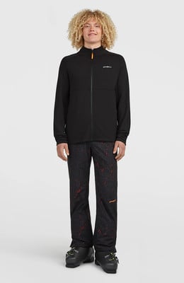 Ein Mann mit lockigem Haar trägt eine schwarze Jacke und gemusterte Hose.