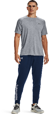 Ein Mann im grauen T-Shirt und dunkelblauer Jogginghose mit Logo steht vor weißem Hintergrund.