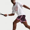 Mann im weißen T-Shirt mit Aufdruck VAMOS und weinroten Shorts spielt Tennis.
