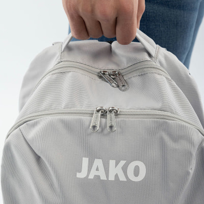 Ein grauer Rucksack mit JAKO-Logo wird von einer Person in Jeans gehalten. Ein grauer Rucksack mit JAKO-Logo wird von einer Person in Jeans gehalten.