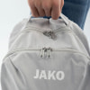Ein grauer Rucksack mit JAKO-Logo wird von einer Person in Jeans gehalten.