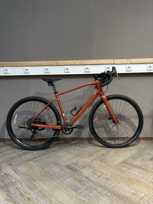 Ein orangefarbenes Gravelbike steht auf einem Holzboden vor einer grauen Wand. Ein orangefarbenes Gravelbike steht auf einem Holzboden vor einer grauen Wand.