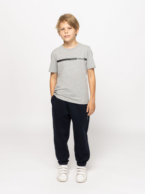 Egy fiú szürke csíkos pólóban és sötét melegítőben áll fehér háttér előtt. Egy fiú szürke csíkos pólóban és sötét melegítőben áll fehér háttér előtt.