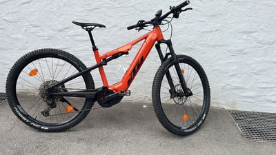 Ein orangefarbenes KTM E-Bike steht vor einer weißen Wand auf Betonboden. Ein orangefarbenes KTM E-Bike steht vor einer weißen Wand auf Betonboden.