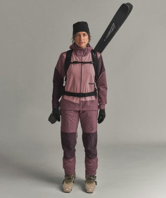 Eine Frau in Skibekleidung mit Skiern auf dem Rücken posiert vor neutralem Hintergrund.