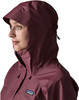Frau in dunkelweinvogelroter Jacke mit Kapuze von der Seite, weißer Hintergrund.