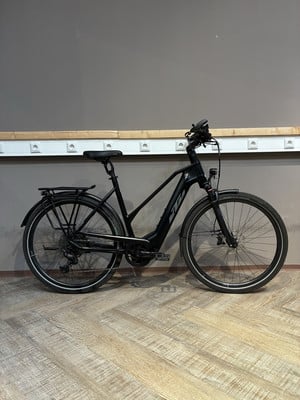 Ein schwarzes KTM E-Bike mit Gepäckträger steht auf einem Holzboden vor einer grauen Wand. Ein schwarzes KTM E-Bike mit Gepäckträger steht auf einem Holzboden vor einer grauen Wand.