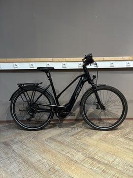 Ein schwarzes KTM E-Bike mit Gepäckträger steht auf einem Holzboden vor einer grauen Wand.