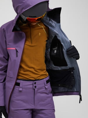 Das Model trägt eine lila Skijacke und Hose, ein orangefarbenes Sweatshirt und eine Skibrille. Das Model trägt eine lila Skijacke und Hose, ein orangefarbenes Sweatshirt und eine Skibrille.