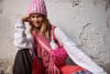 Eine Frau in rosa Mütze und Schal mit Handtasche sitzt an einer Wand.