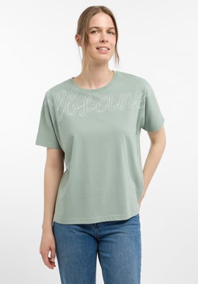Eine Frau in einem grünen T-Shirt mit Aufdruck und blauer Jeans posiert vor weißem Hintergrund.