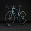 Modernes Gravelbike mit blaugrünem Rahmen und braunen Reifen vor dunklem Hintergrund.