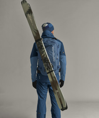 Ein Mann in blauer Kleidung trägt Skier auf dem Rücken.