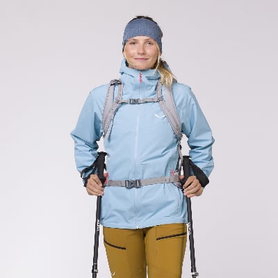 Eine Frau in einer blauen Jacke und braunen Hose hält Wanderstöcke.