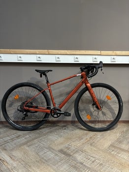 Ein orangefarbenes Gravelbike steht auf einem Holzboden vor einer grauen Wand.