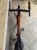 Orangefarbenes Gravelbike mit schwarzem Lenker und Sattel auf Holzboden.