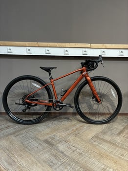 Ein orangefarbenes Gravelbike steht auf einem Holzboden vor einer grauen Wand.