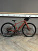 Ein orangefarbenes Gravelbike steht auf einem Holzboden vor einer grauen Wand.