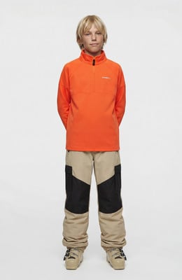 Junge in einem orangefarbenen Pullover und einer beigen Hose mit schwarzen Einsätzen.