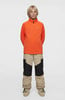 Junge in einem orangefarbenen Pullover und einer beigen Hose mit schwarzen Einsätzen.