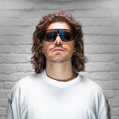 Ein Mann mit lockigem Haar und Sonnenbrille vor einer weißen Ziegelwand. Ein Mann mit lockigem Haar und Sonnenbrille vor einer weißen Ziegelwand.