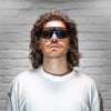 Ein Mann mit lockigem Haar und Sonnenbrille vor einer weißen Ziegelwand.