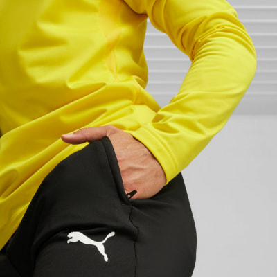 Ein Mann im gelben Pullover steckt seine Hand in die Tasche einer schwarzen Hose mit Puma-Logo. Ein Mann im gelben Pullover steckt seine Hand in die Tasche einer schwarzen Hose mit Puma-Logo.