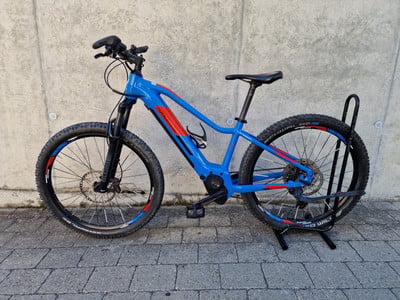 Ein blaues Mountain-E-Bike mit roten Akzenten auf einem Ständer vor einer Wand. Ein blaues Mountain-E-Bike mit roten Akzenten auf einem Ständer vor einer Wand.