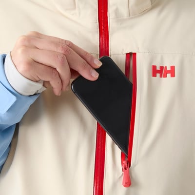 Eine Hand steckt ein Handy in eine Jackentasche mit rotem Reißverschluss und HH-Logo. Eine Hand steckt ein Handy in eine Jackentasche mit rotem Reißverschluss und HH-Logo.