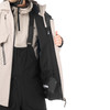 Ein Mann in einer beigen Jacke und schwarzer Latzhose mit offener Jacke.