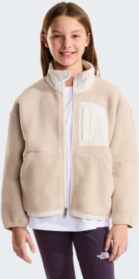 Ein Mädchen in einer beige Fleecejacke mit weißem T-Shirt und lila Hose.