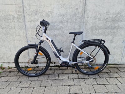 Ein silbernes E-Bike mit Gepäckträger steht vor einer Betonwand.