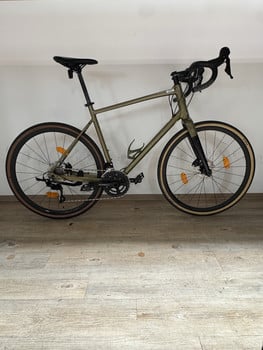 Ein grünes Gravelbike steht auf einem Holzboden vor einer weißen Wand.