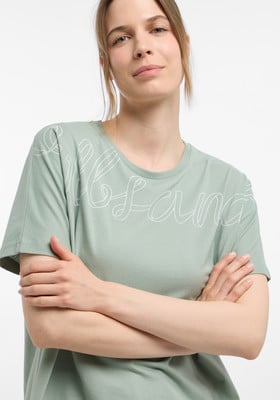 Eine Frau in einem grünen T-Shirt mit weißer Aufschrift und verschränkten Armen.