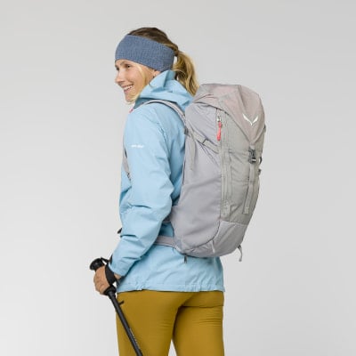 Frau mit Rucksack und Wanderstöcken vor grauem Hintergrund.